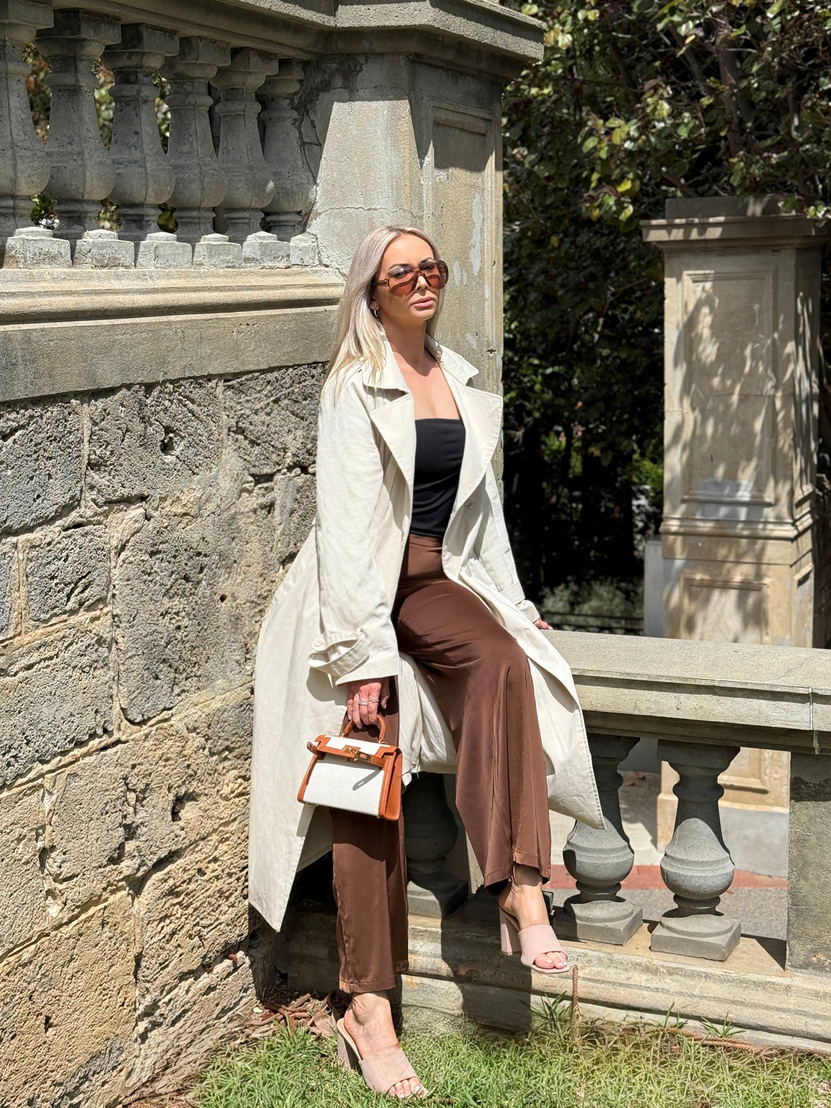 The Amélie trench coat in stone, full-length seated view