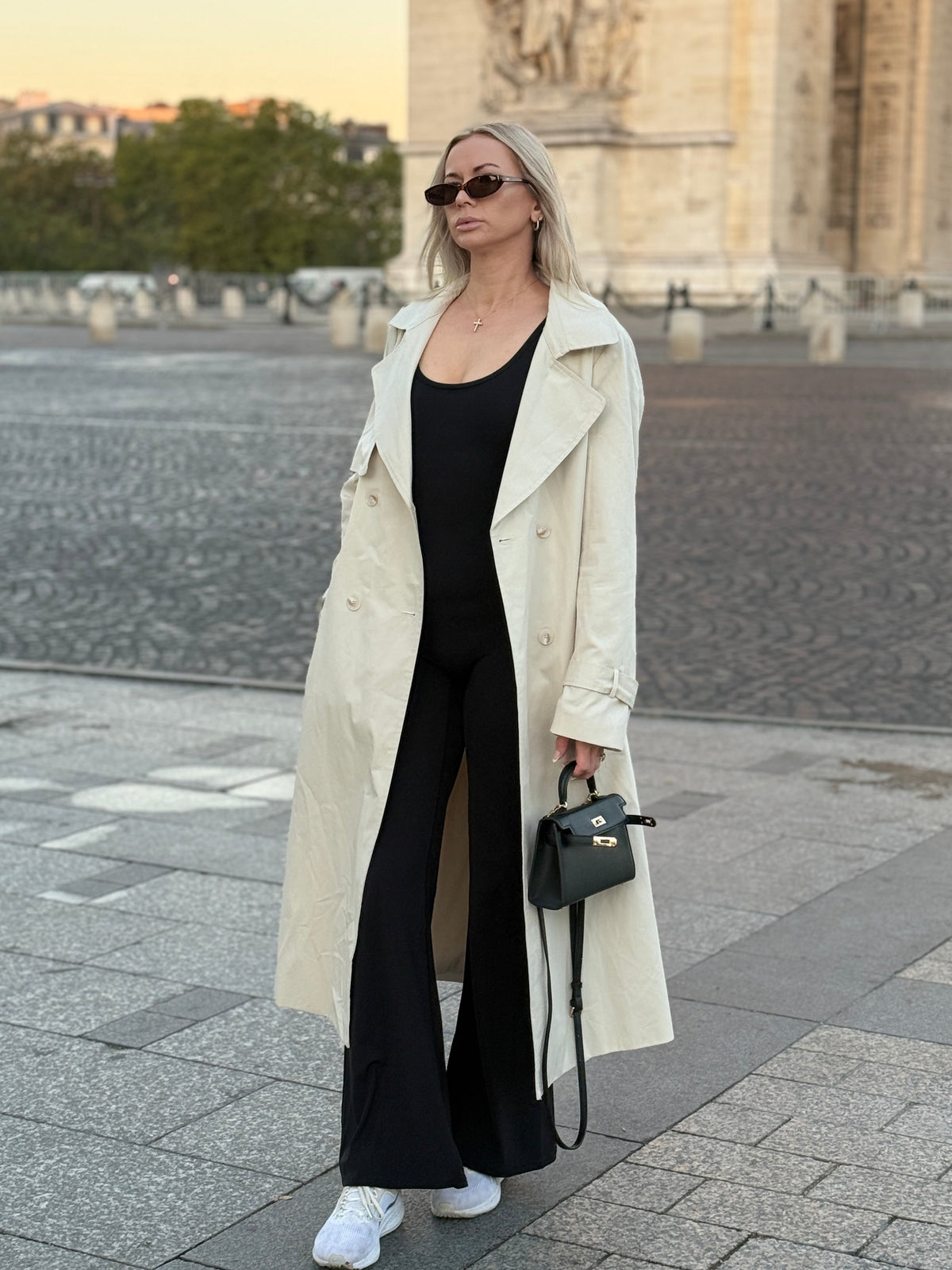 The Amélie trench coat in stone, full-length front view beside a stone arch