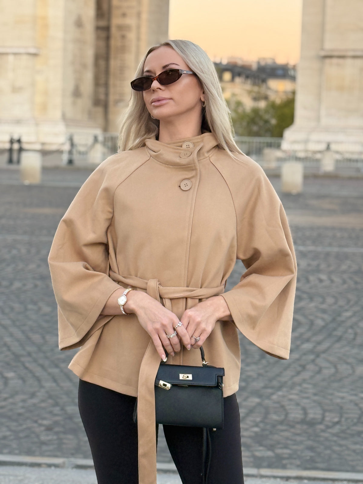 The Éloise cape coat with high neckline and waist tie, front view beside a stone arch