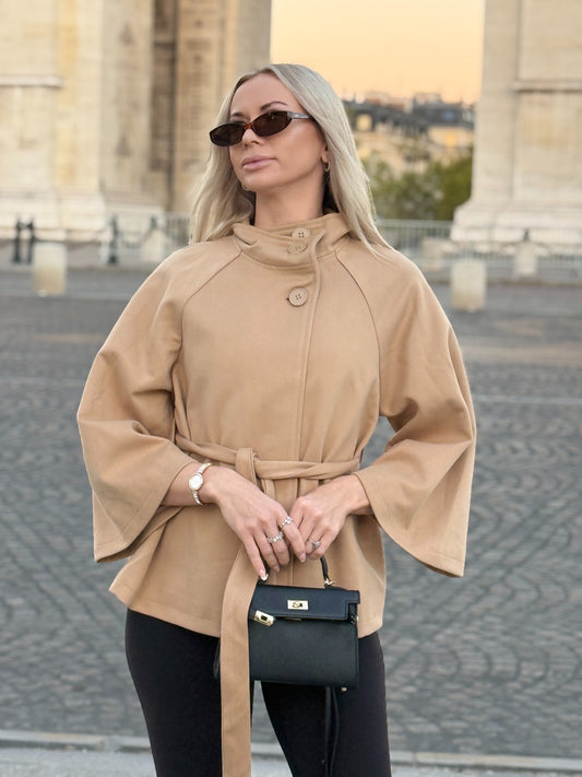 The Éloise cape coat with high neckline and waist tie, front view beside a stone arch